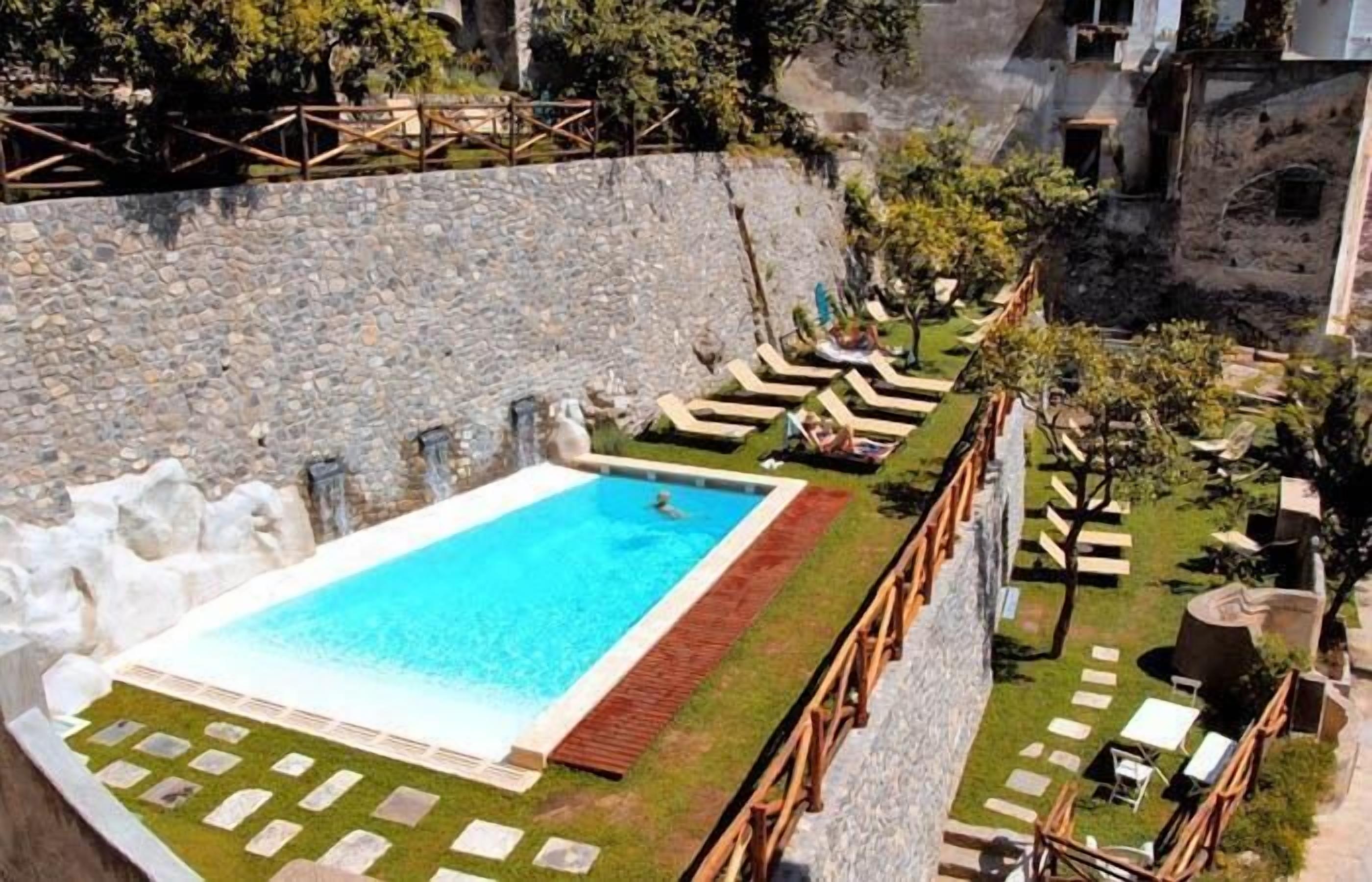 A swimming pool with a wooden deck and lounge chairs, surrounded by a stone wall and greenery.