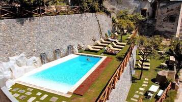 A swimming pool with a wooden deck and lounge chairs, surrounded by a stone wall and greenery.