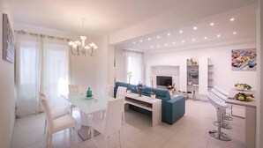 A modern living room with a blue sofa, white dining table, and chairs. There is a TV, a bookshelf, and a chandelier. - Modern Apartment close to the Beach - Corso 4 by AMALFIVACATION.IT (MINORI)