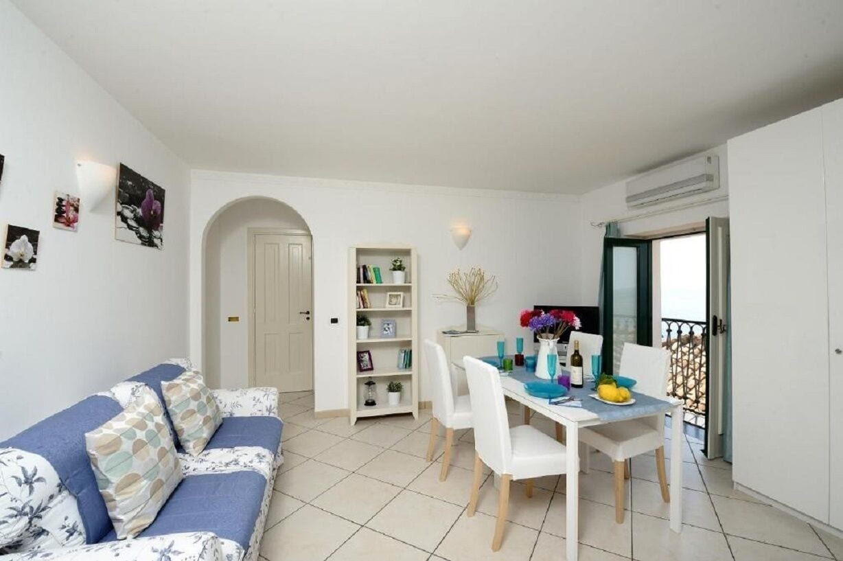 Een knus woongedeelte met een bank, eettafel, stoelen, een boekenkast en een klein balkon.