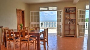Un coin repas avec une table et des chaises en bois, une vue sur la mer par des portes ouvertes et une étagère ornée d’objets décoratifs.