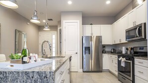 A modern kitchen with white cabinets, stainless steel appliances, granite countertops, and pendant lighting. - Disney Getaway – 5 Min Drive, Sleeps 8 (Kissimmee)