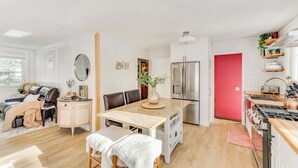 Eine moderne Küche mit Kochinsel, ein Essbereich mit Tisch und Stühlen, ein Wohnzimmer mit Sofa und Couchtisch und eine rote Tür.