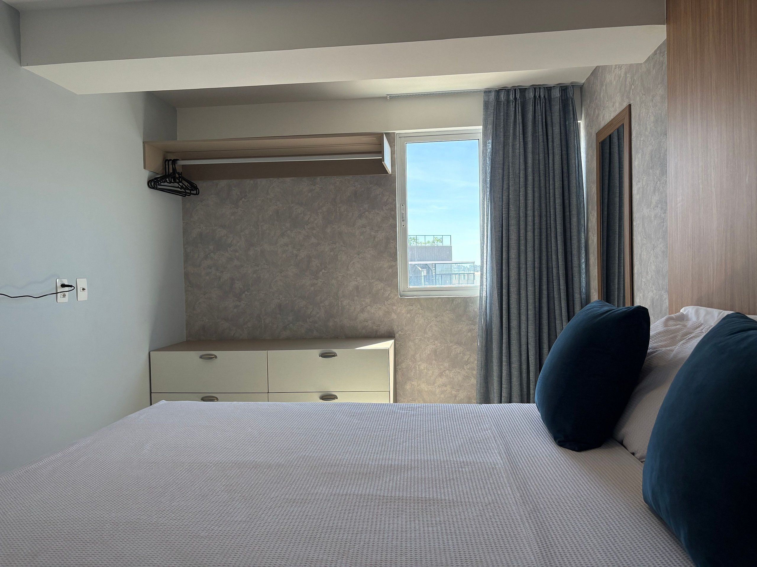 Quarto com cama, cómoda, janela com cortinas e candeeiro fixo na parede.