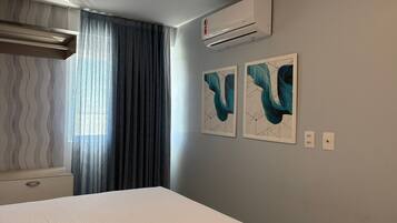 A hotel room with a bed, air conditioning unit, curtains, and two abstract paintings on the wall.