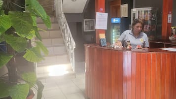 Una recepción de hotel con un letrero digital, una planta grande en maceta y una escalera al fondo.