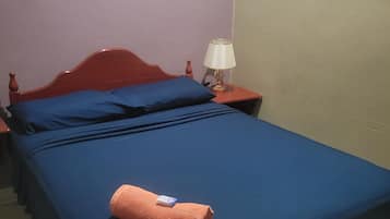 Una cama con ropa de cama azul y una toalla enrollada, una mesita de noche con lámpara, y un cuadro en la pared.