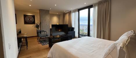 A modern hotel room with a large bed, a TV, a seating area, and a view of the city.