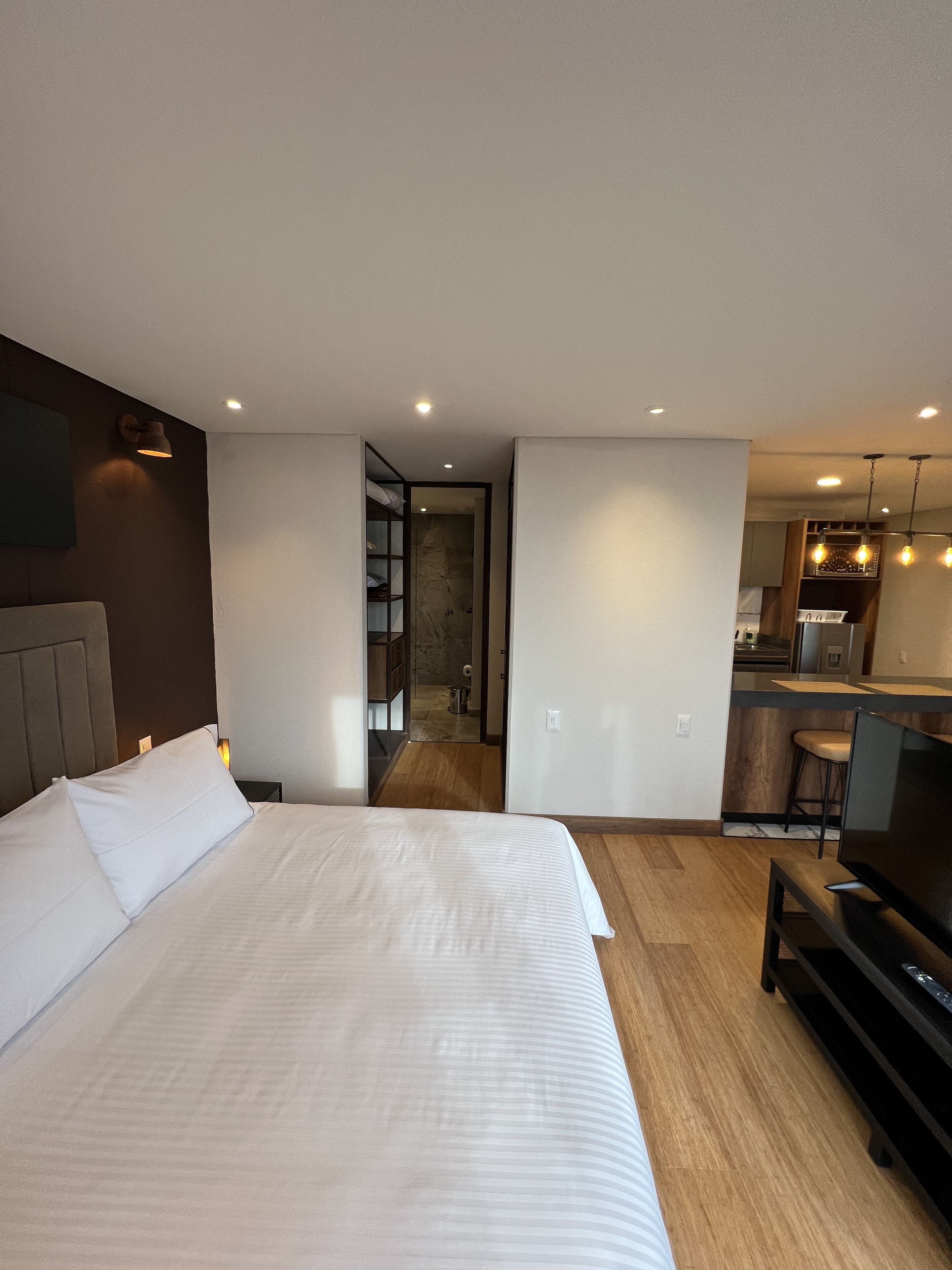 Habitació d'hotel moderna amb un llit gran, televisió, petita cuina i un bany visible a través d'una porta oberta.
