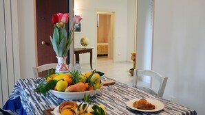 Une table à manger recouverte d’une nappe à motifs, un saladier rempli de fruits, une assiette avec un croissant et un vase de fleurs.