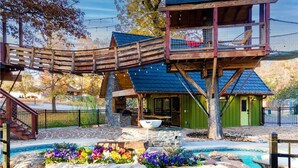 A rustic cabin with a wooden deck, a stone fireplace, and a swimming pool surrounded by colorful flowers.