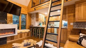 A cozy cabin interior with a fireplace, wooden walls, a kitchen area, and a loft space.