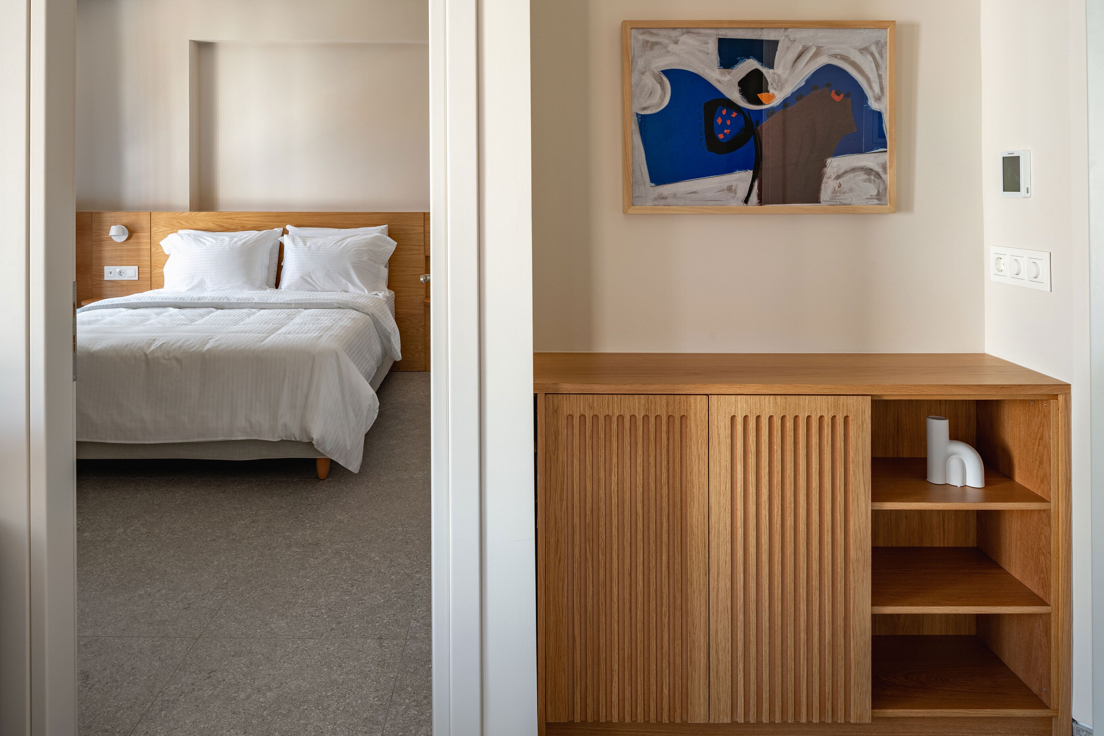 A hotel room with a bed, a wooden cabinet with shelves, and a painting on the wall.