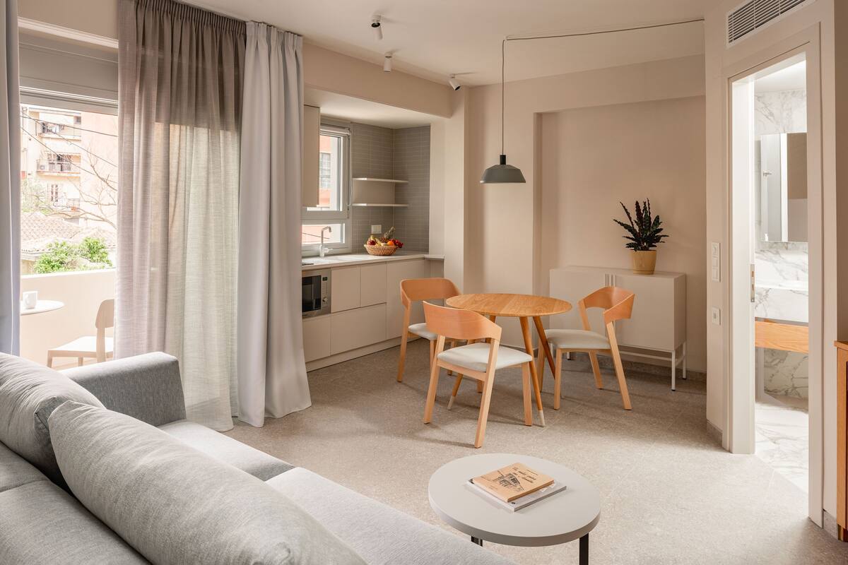 Una sala moderna con un sofá gris, una mesa de comedor redonda con sillas de madera, y una pequeña mesa redonda con un libro.
