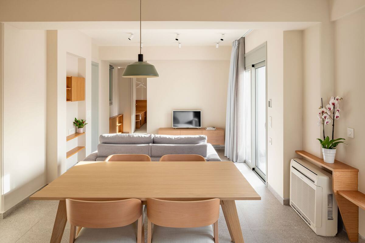 Un comedor moderno con una mesa y sillas de madera, una lámpara colgante y vistas a una sala de estar con un sofá y televisor.