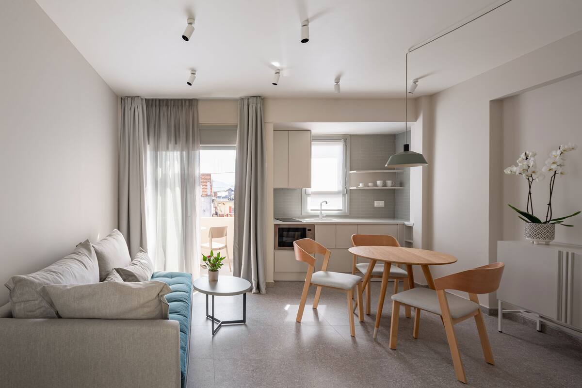 Una sala moderna con un sofá, una mesita y un comedor con sillas. Se observa una cocina al fondo con armarios y una ventana.