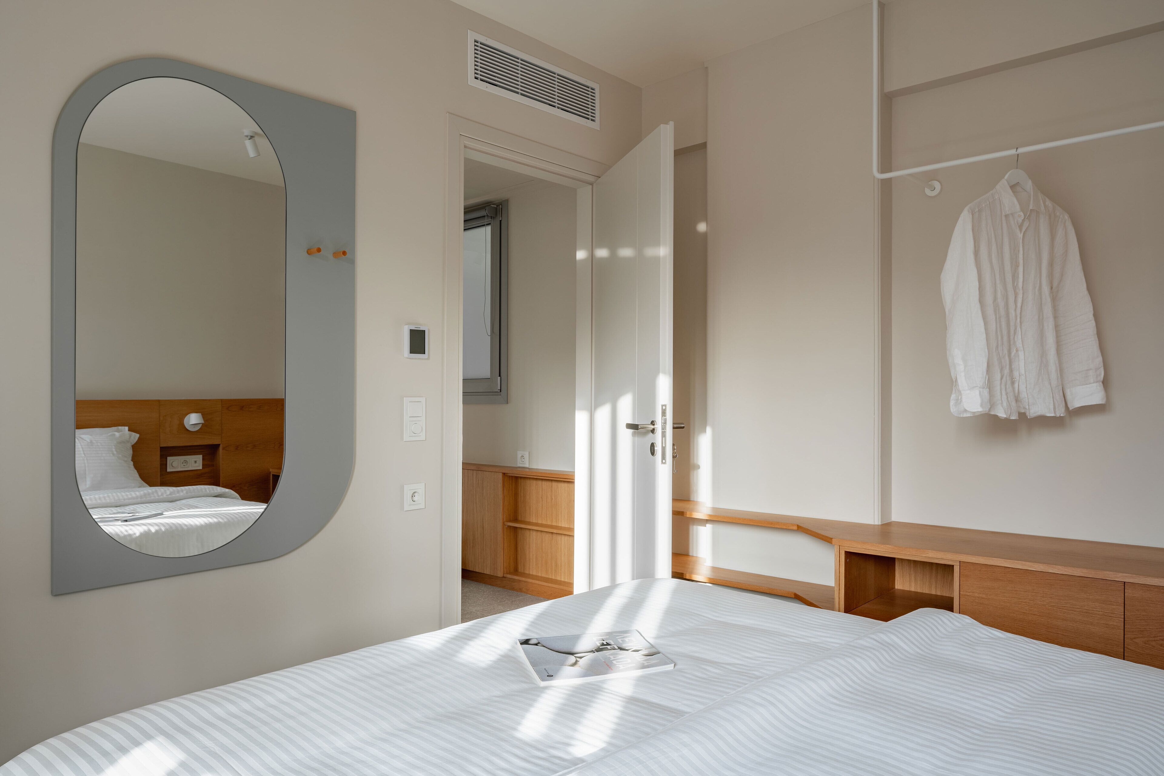Une chambre d’hôtel avec un lit, un miroir, une table de chevet en bois, une robe suspendue et une vue sur la salle de bain.