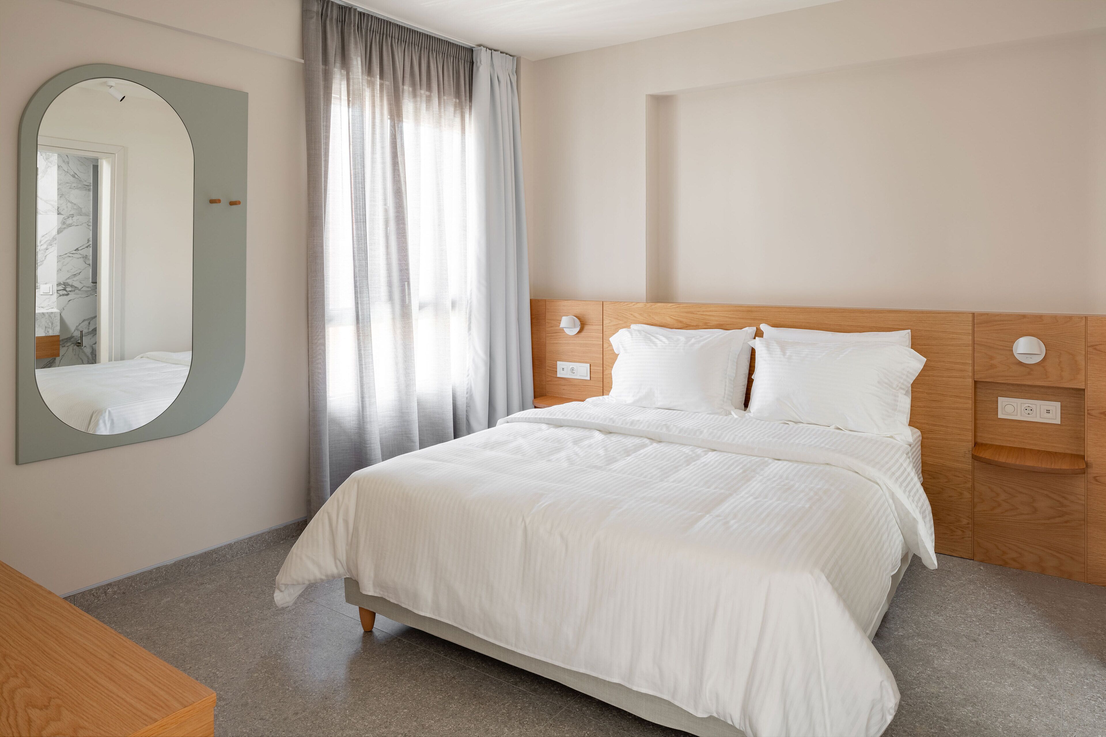 A neatly arranged hotel room with a large bed, a wooden nightstand, a mirror, and a window with curtains.