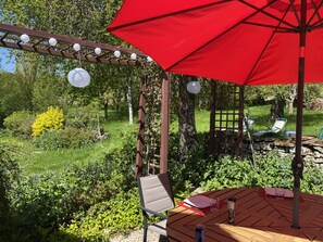 Uma mesa de madeira com um guarda-sol vermelho, rodeada por vegetação e uma pérgula com lanternas penduradas.