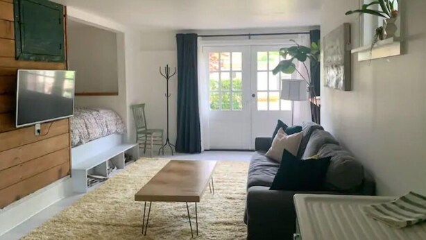 Un salon comprenant un canapé, une table basse, un téléviseur fixé à un mur en bois, un fauteuil et une grande fenêtre avec des rideaux.