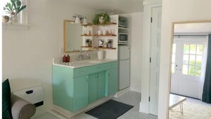 Une salle de bain avec un meuble-lavabo de couleur turquoise, un lavabo blanc et un grand miroir. Des étagères sont placées au-dessus du meuble-lavabo et contiennent divers objets. On peut également apercevoir un four à micro-ondes et un réfrigérateur à l’arrière-plan.