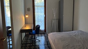 Une petite pièce bien éclairée, comprenant un lit, un bureau avec une chaise, un lampadaire et une armoire.