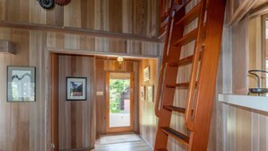 Un couloir intérieur en bois, avec une échelle en bois, des tableaux encadrés accrochés aux murs et une porte donnant sur l’extérieur.