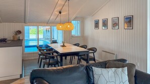 A modern living room with a black leather sofa, a dining table with chairs, a large window overlooking a pool, and framed pictures on the wall. - Awesome home in Ringkøbing with sauna (Ringkøbing)