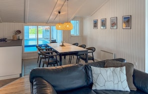 A modern living room with a black leather sofa, a dining table with chairs, a large window overlooking a pool, and framed pictures on the wall.