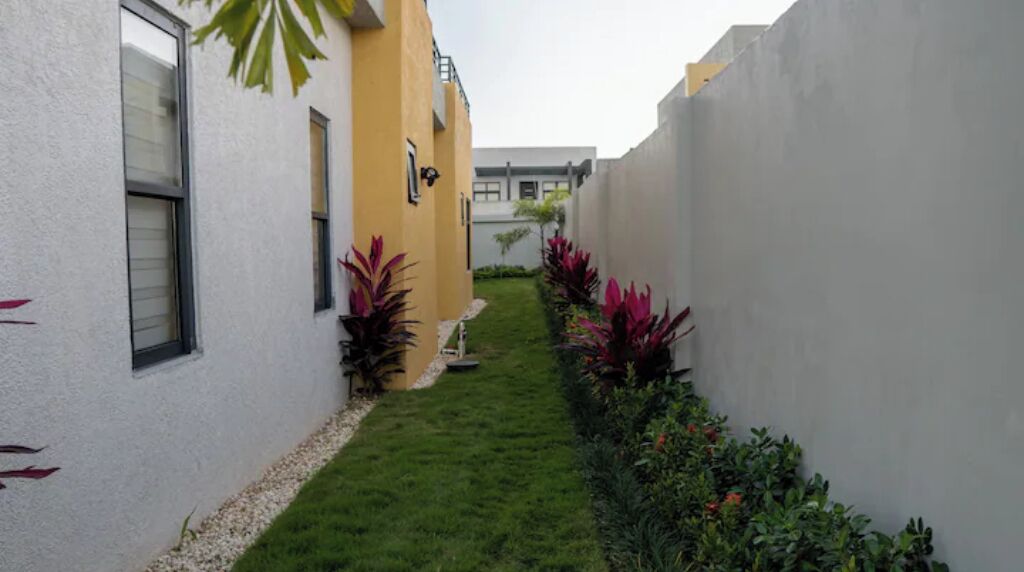 Un étroit sentier bordé de bâtiments aux murs blancs et jaunes, qui mène à un petit espace verdoyant planté d’arbustes.