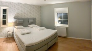 En pænt redt seng med puder og en sengetavlelampe, et vindue med persienner og en radiator på væggen.