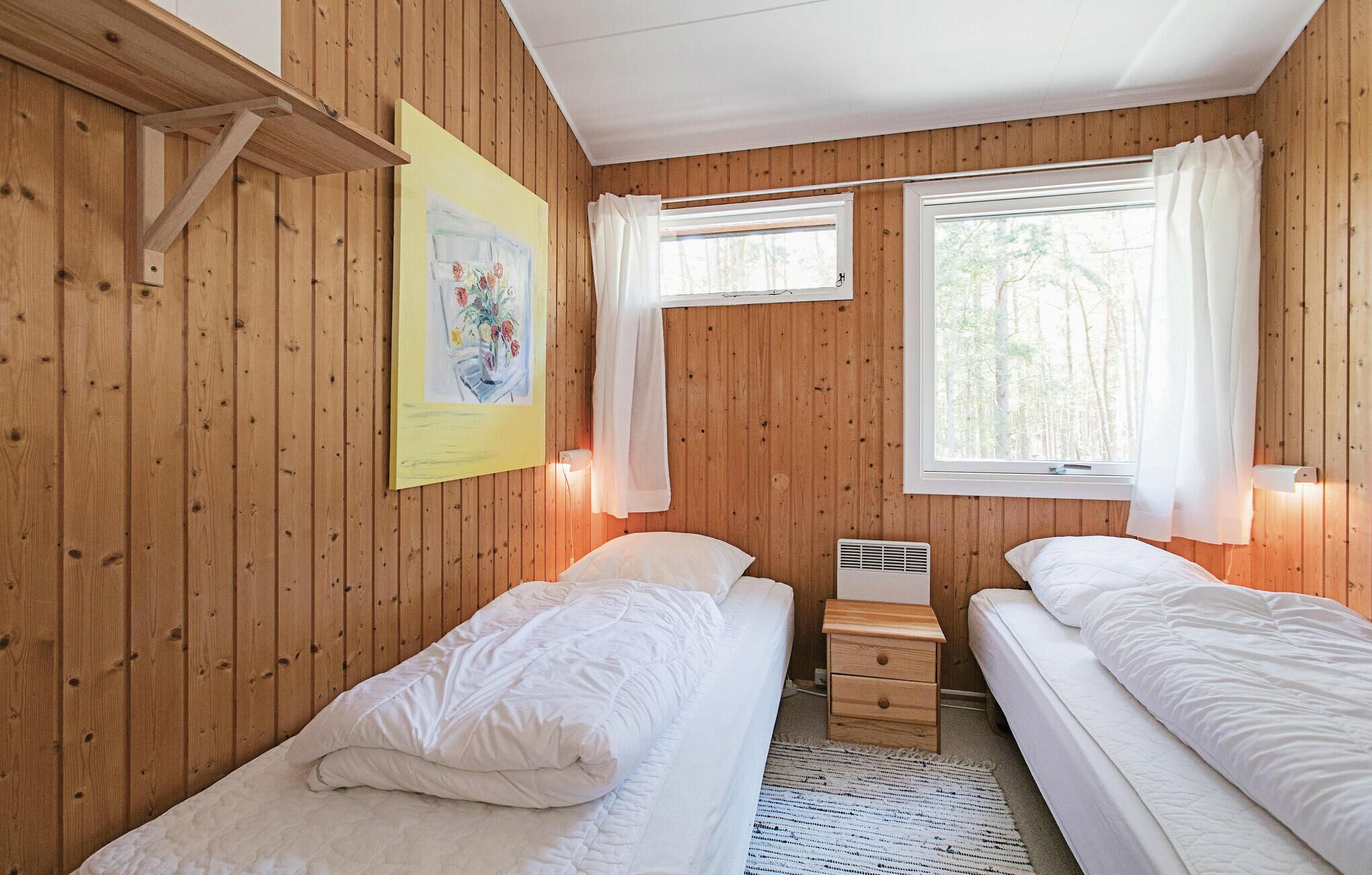 A room with wooden walls, two beds, a small bedside table, a radiator, and a window with curtains.