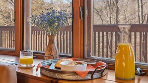 Un plateau de petit-déjeuner avec des toasts, des céréales et un verre de jus d’orange, posé sur une table en bois près d’une fenêtre donnant sur une terrasse en bois et des arbres.