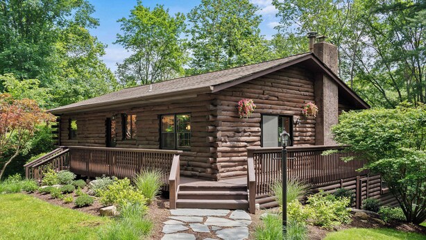 Une cabane en rondins avec un chemin de pierres menant à l’entrée, entourée d’une végétation luxuriante et dotée d’une terrasse en bois.