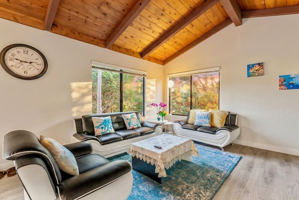 A modern living room with a wooden ceiling, two sofas, a coffee table, and large windows with a view of trees.