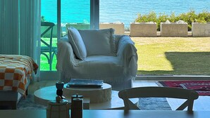 A room with a large window overlooking the sea, a white armchair, a round table, and a bed with an orange and white checkered bedspread. - Henley Beachfront Luxury Home With Private Pool, Spa And Sauna! (Henley Beach South)