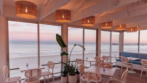 Ein Restaurant direkt am Strand mit großen Fenstern, Korbmöbeln und hängenden Lampen aus Korb.