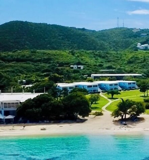A coastal property with a beach, a large building with multiple windows, and a hilly landscape in the background.
