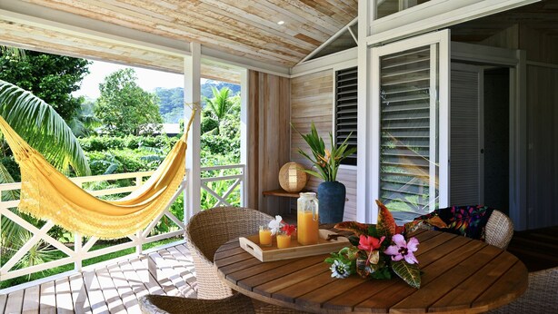 A wooden deck with a hammock, wicker furniture, a table with drinks and flowers, and a view of greenery. - FARE VAVAE (Raiatea)