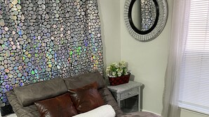 A bedroom with a grey sofa bed, a decorative mirror, a potted plant, and a tiled floor. - Delightful, 4BR 2.5BA ranch style near Parks, trails, shops, malls, Restaurants. (Lithonia)