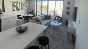A modern kitchen with a white countertop, black stools, and a dining area with a round table and chairs. There is a large window with a view of buildings and a balcony. - Apartment in Sarasota (Sarasota)