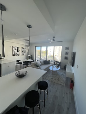 A modern kitchen with a white countertop, black stools, and a dining area with a round table and chairs. There is a large window with a view of buildings and a balcony. - Apartment in Sarasota (Sarasota)