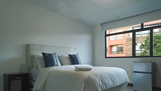 A bedroom with a bed, a nightstand, a refrigerator, and a window with a view of buildings and trees. - Privated room with bathroom (Bogotá)
