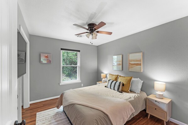 A bedroom with a bed, nightstands, a ceiling fan, a window, and artwork on the walls.