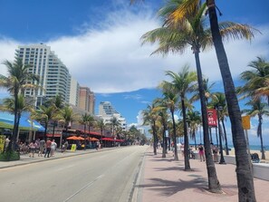 Une rue bordée de palmiers et de restaurants situés en bord de mer, avec un ciel dégagé et une vue sur l’océan.
