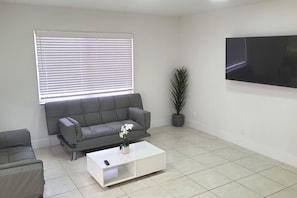 A living room with a grey sofa, a white coffee table, a potted plant, and a flat-screen TV mounted on the wall.