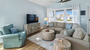 A living room with a sofa, armchair, TV, and a view of the outside. - WATERSHED COTTAGE, MODERN 3BD/2BA HOME W/POOL & FIRE PIT! PET FRIENDLY! (Mexico Beach)