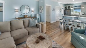 A modern living room with a beige sofa, a round coffee table, and a dining area with white chairs and a kitchen counter. - WATERSHED COTTAGE, MODERN 3BD/2BA HOME W/POOL & FIRE PIT! PET FRIENDLY! (Mexico Beach)