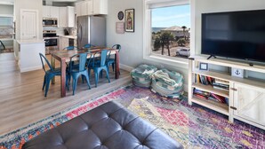 Una cocina moderna con armarios blancos, una zona de comedor con una mesa de madera y sillas azules, un televisor grande sobre un mueble de madera, y vistas del exterior a través de una ventana.