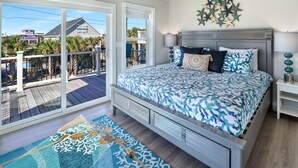 Un dormitorio con una cama grande, un ventanal corredero a un balcón y vistas a casas y palmeras.